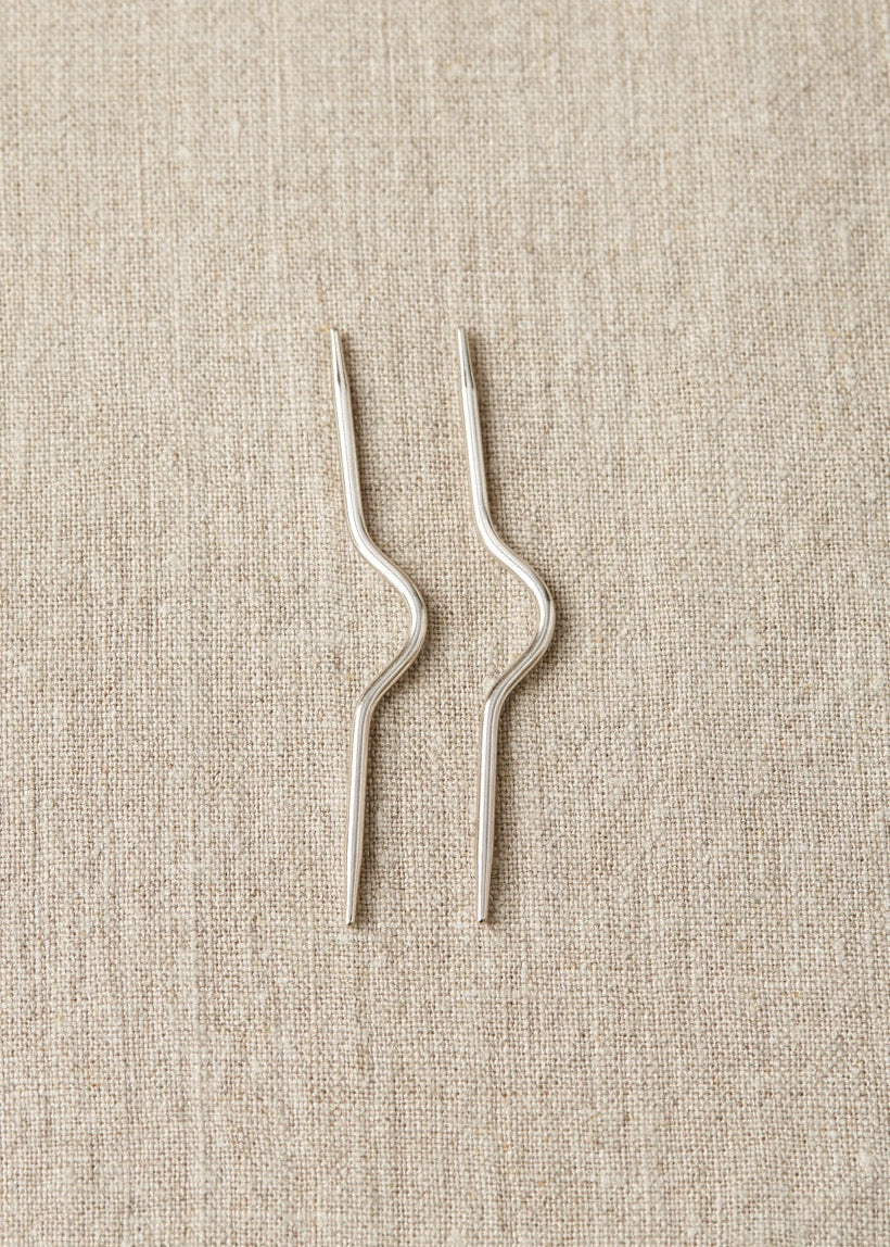 Two nickel plated steel curved cable knitting needles placed on a textured fabric surface.