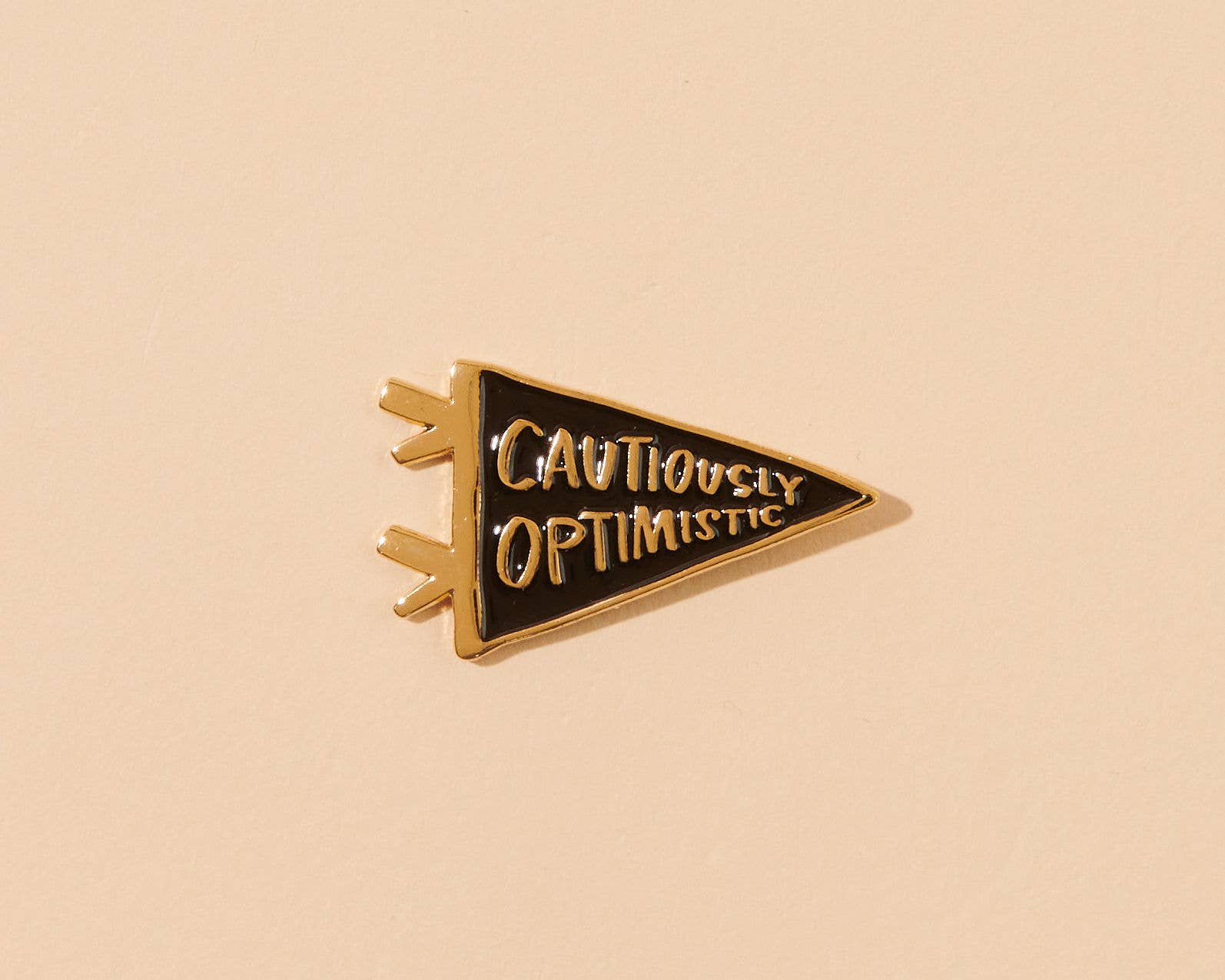Triangular enamel pin with 'CAUTIOUSLY OPTIMISTIC' text on a beige background